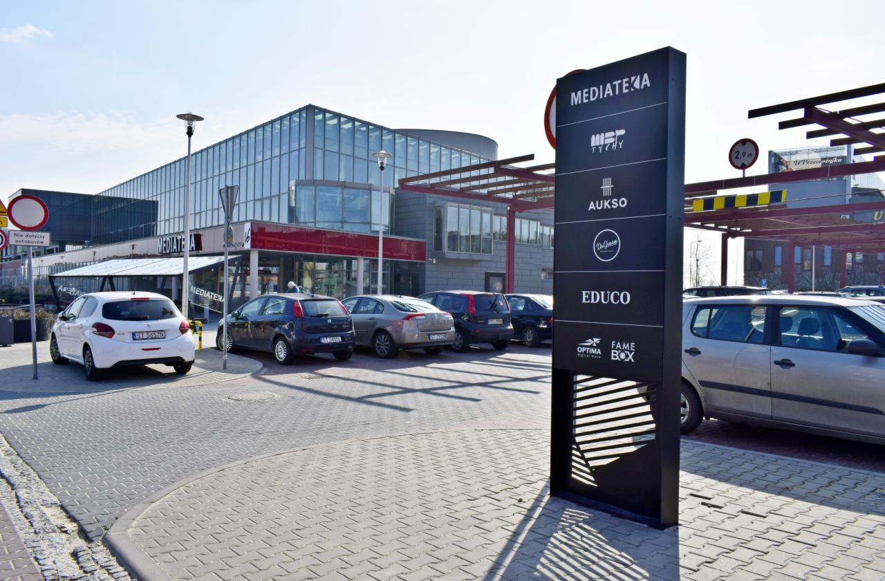 Zewnętrzny pylon reklamowy z logotypami firm przed nowoczesnym budynkiem z przeszkloną fasadą i parkingiem, w tle widoczne znaki drogowe.