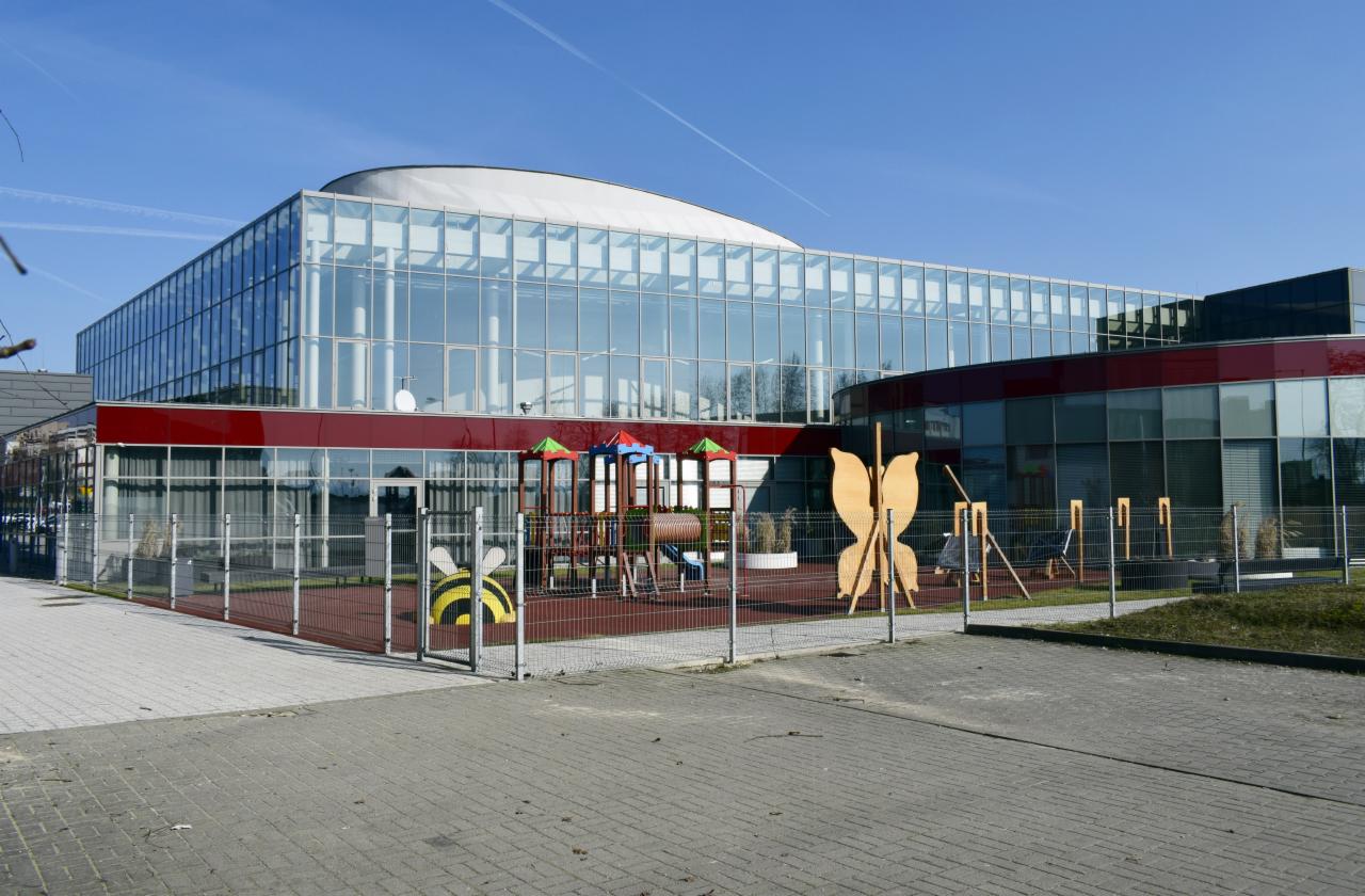 Nowoczesny budynek z przeszkleniami i czerwonym pasem w Tychach, otoczony placem zabaw z drewnianą motylem i pszczołą na ogrodzeniu.