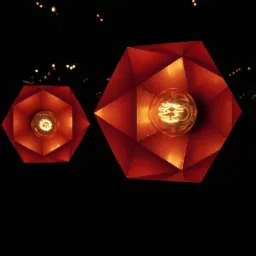 Dwie geometryczne, metalowe lampy w odcieniach czerwieni i pomarańczy wiszące na czarnym tle, z widocznymi żarówkami edisona i drobnymi światełkami w tle.