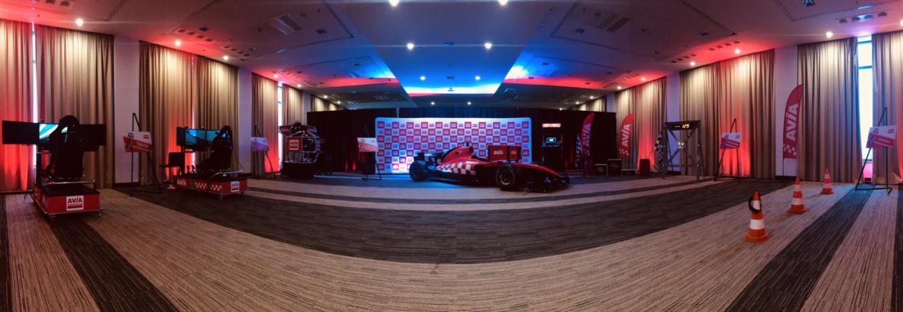 Sala konferencyjna z czerwonym samochodem wyścigowym na podeście, symulatory wyścigowe po bokach, czerwone i niebieskie oświetlenie sceniczne, flagi i pachołki.