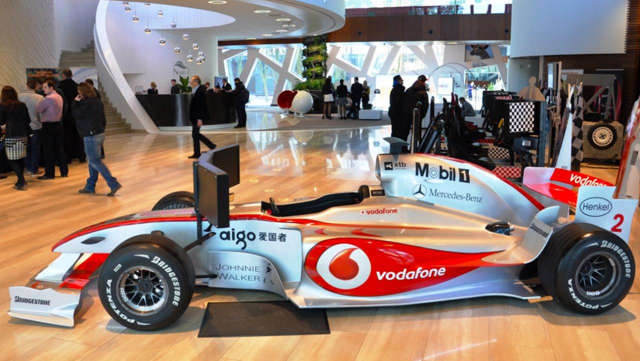 Bolid Formuły 1 McLaren Vodafone zaparkowany w nowoczesnym, przeszklonym wnętrzu budynku z widocznymi schodami, recepcją i przechodzącymi osobami.