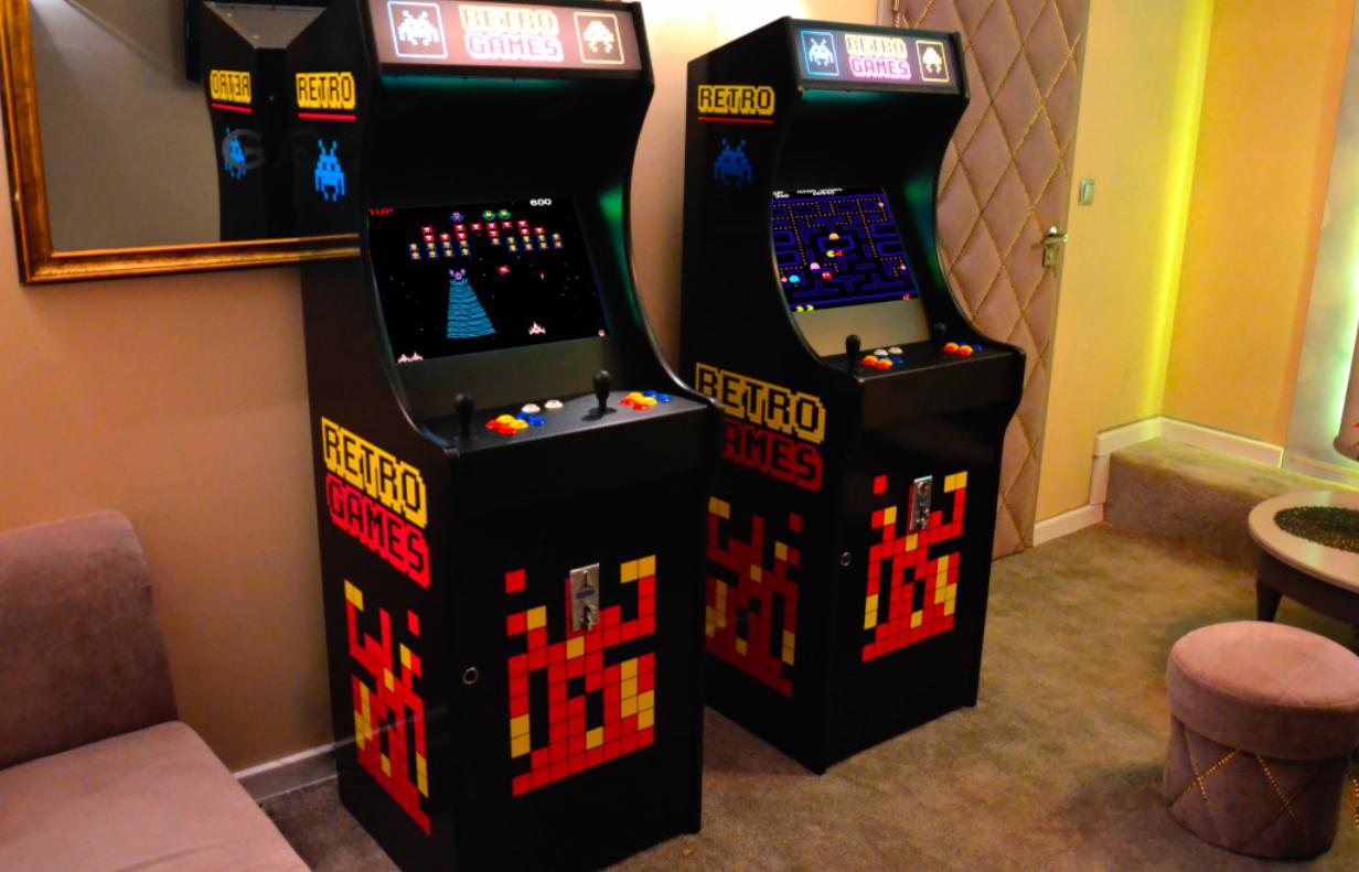 Dwa automaty do gier retro z motywem Space Invaders i Pac-Man, ustawione w eleganckim wnętrzu z pikowanymi ścianami i złotym lustrem.