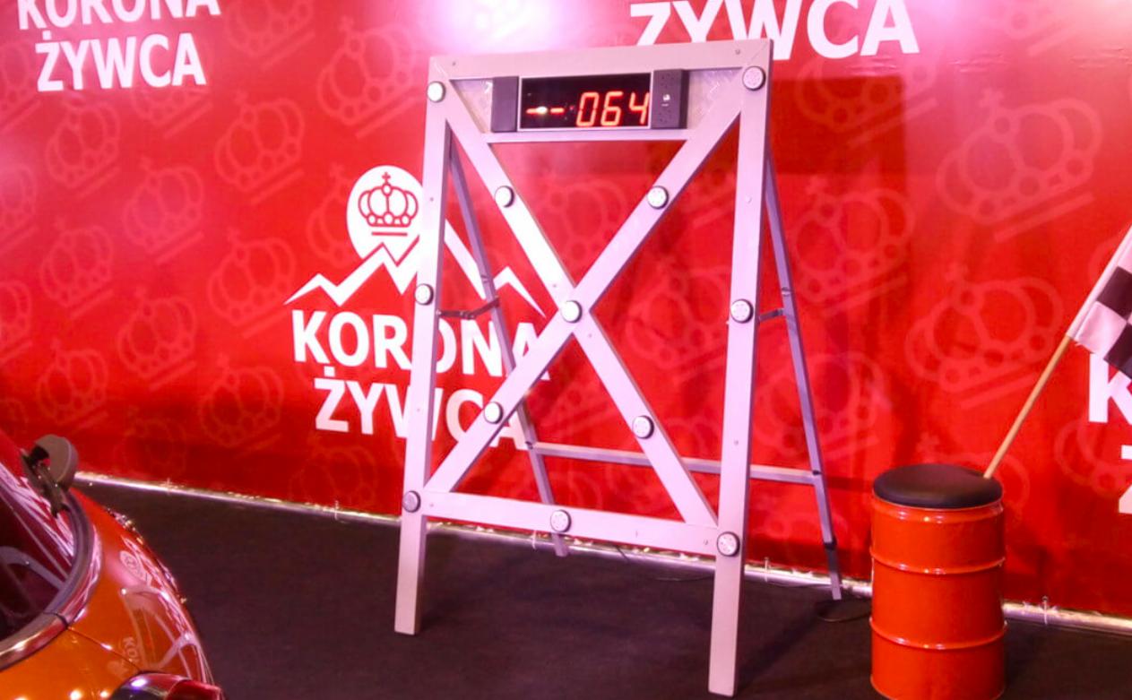 Metalowa konstrukcja z wyświetlaczem z numerem 064, stojąca obok czerwonego stołka imitującego beczkę i flagi w szachownicę, na tle czerwonej ściany z logo Korona Żywca.