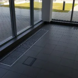 Wnętrze z szarymi płytkami podłogowymi i kanałami grzewczymi pod oknami.