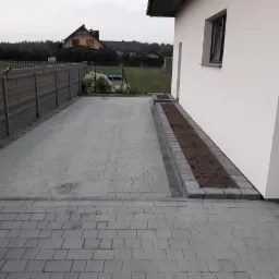 Szara nawierzchnia z kostki brukowej prowadząca do domu z przygotowanym miejscem na rabatę kwiatową wzdłuż ściany i ogrodzeniem w tle.