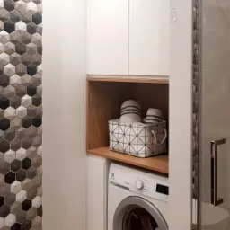 Nowoczesna łazienka z pralką zabudowaną w szafce, nad nią półka z koszem na pranie, ściana wyłożona płytkami heksagonalnymi w odcieniach szarości i bieli.