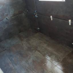 Renova Remonty i Wykończenia Wnętrz - Układanie ciemnych płytek imitujących metal w narożnej wnęce, z widocznymi krzyżykami dystansowymi i klinami poziomującymi.