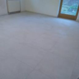 Renova Remonty i Wykończenia Wnętrz - Wykończona podłoga z jasnych, kwadratowych płytek w pustym pokoju z grzejnikiem i oknem balkonowym.