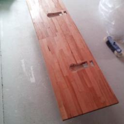 Renova Remonty i Wykończenia Wnętrz - Blat drewniany z wyciętymi otworami leżący na podłodze w trakcie prac wykończeniowych, widoczna szczotka i pojemnik z narzędziami.