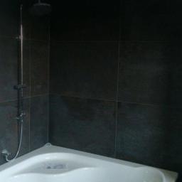 Renova Remonty i Wykończenia Wnętrz - Narożna wanna z ciemnymi płytkami na ścianach i chromowaną deszczownicą w rogu łazienki.