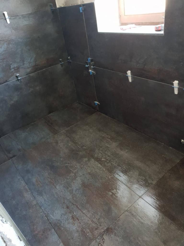 Układanie ciemnych płytek imitujących metal w narożnej wnęce, z widocznymi krzyżykami dystansowymi i klinami poziomującymi.