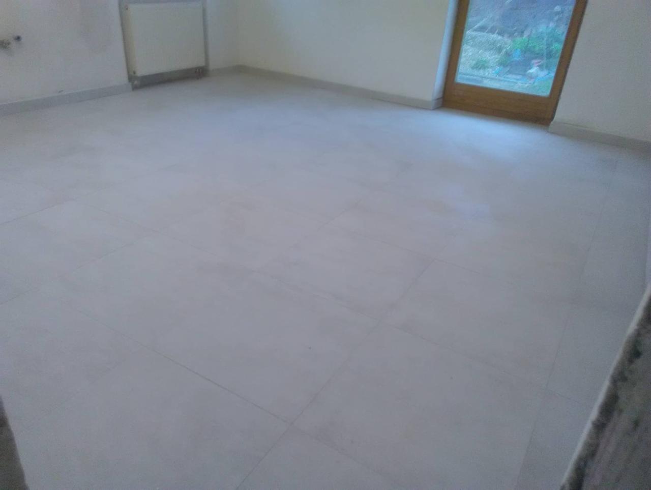 Wykończona podłoga z jasnych, kwadratowych płytek w pustym pokoju z grzejnikiem i oknem balkonowym.