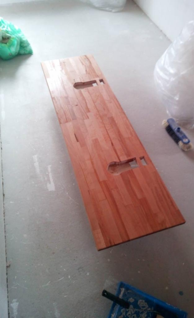 Blat drewniany z wyciętymi otworami leżący na podłodze w trakcie prac wykończeniowych, widoczna szczotka i pojemnik z narzędziami.