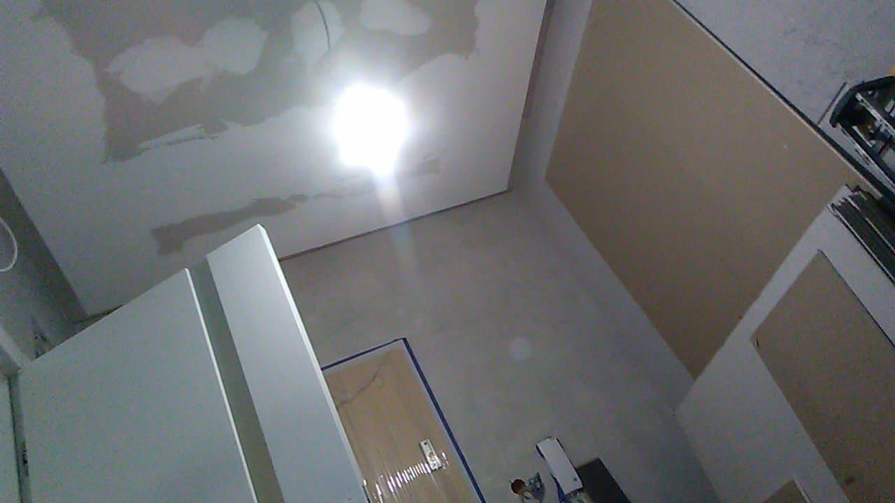 Niedokończona powierzchnia sufitu ze śladami świeżo nałożonej szpachli, widoczny fragment ściany z płytą karton-gips, drzwi oklejone taśmą malarską i narzędzia malarskie w rogu pomieszczenia.