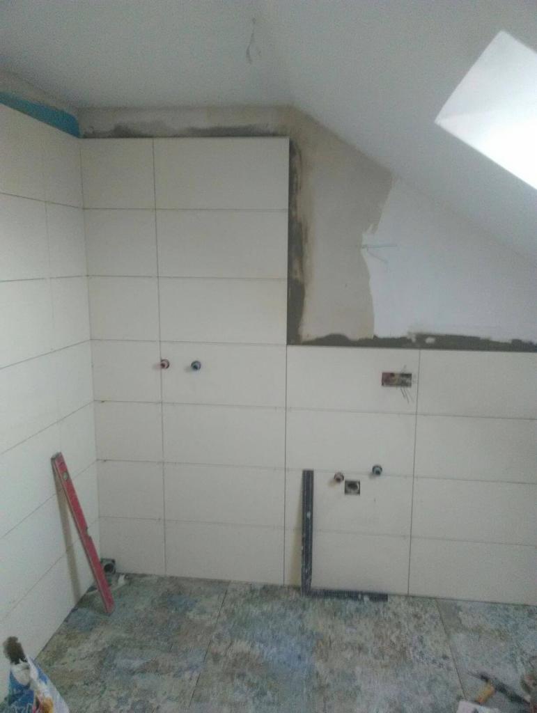 Układanie jasnych płytek w łazience na poddaszu; widoczne niedokończone fragmenty ściany, poziomica i kątownik na podłodze.