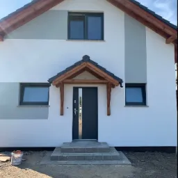 Nowoczesny dom jednorodzinny z białoszarą elewacją, drewnianym zadaszeniem nad wejściem i trzema schodami wejściowymi, widok z przodu, w tle błękitne niebo.