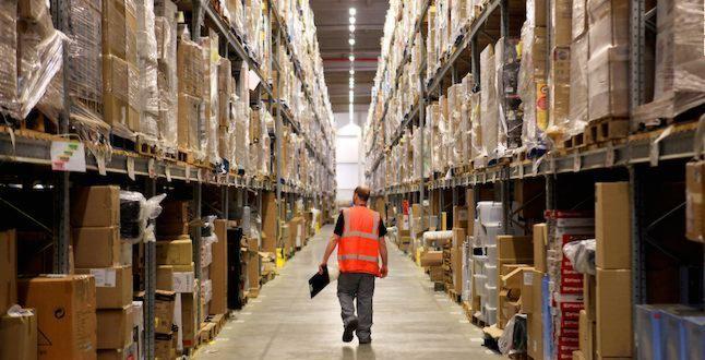 Pracownik w pomarańczowej kamizelce idzie korytarzem magazynu pełnym regałów z zapakowanymi towarami.