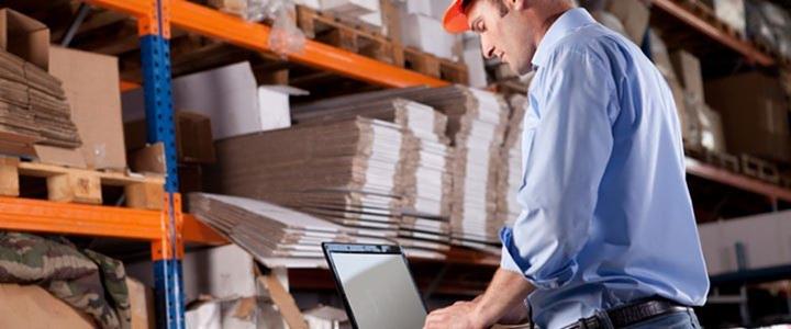 Pracownik w kasku wprowadza dane do laptopa w magazynie z regałami wypełnionymi kartonami i paletami.