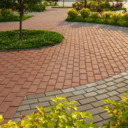 Ścieżka z czerwonej kostki brukowej obramowana szarą kostką, z okrągłym klombem z trawą wokół drzewa i rabatami kwiatowymi w tle.