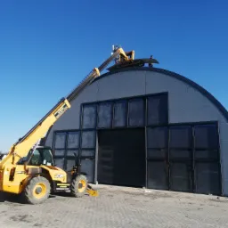 Żółta ładowarka teleskopowa podnosi blachę trapezową na dach hali o konstrukcji łukowej z szarej blachy, pracownik na dachu, błękitne niebo.