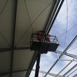 Silverhouse - Montaż paneli warstwowych na konstrukcji stalowej hali z użyciem podnośnika koszowego. Widoczne fragmenty nieba i stalowych elementów konstrukcji.