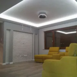 Jasne wnętrze salonu z podświetlanym sufitem kasetonowym LED, szarymi ścianami, imitacją białej cegły na ścianie, drewnianą podłogą i żółtymi meblami wypoczynkowymi.