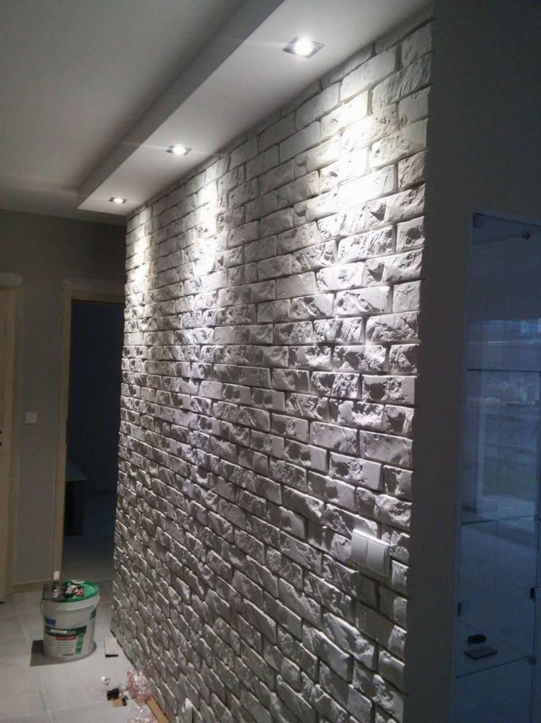 Ściana wykończona dekoracyjną, białą cegłą z widocznymi punktowymi lampami sufitowymi i fragmentem pomieszczenia z drzwiami oraz wiadrem farby w rogu.