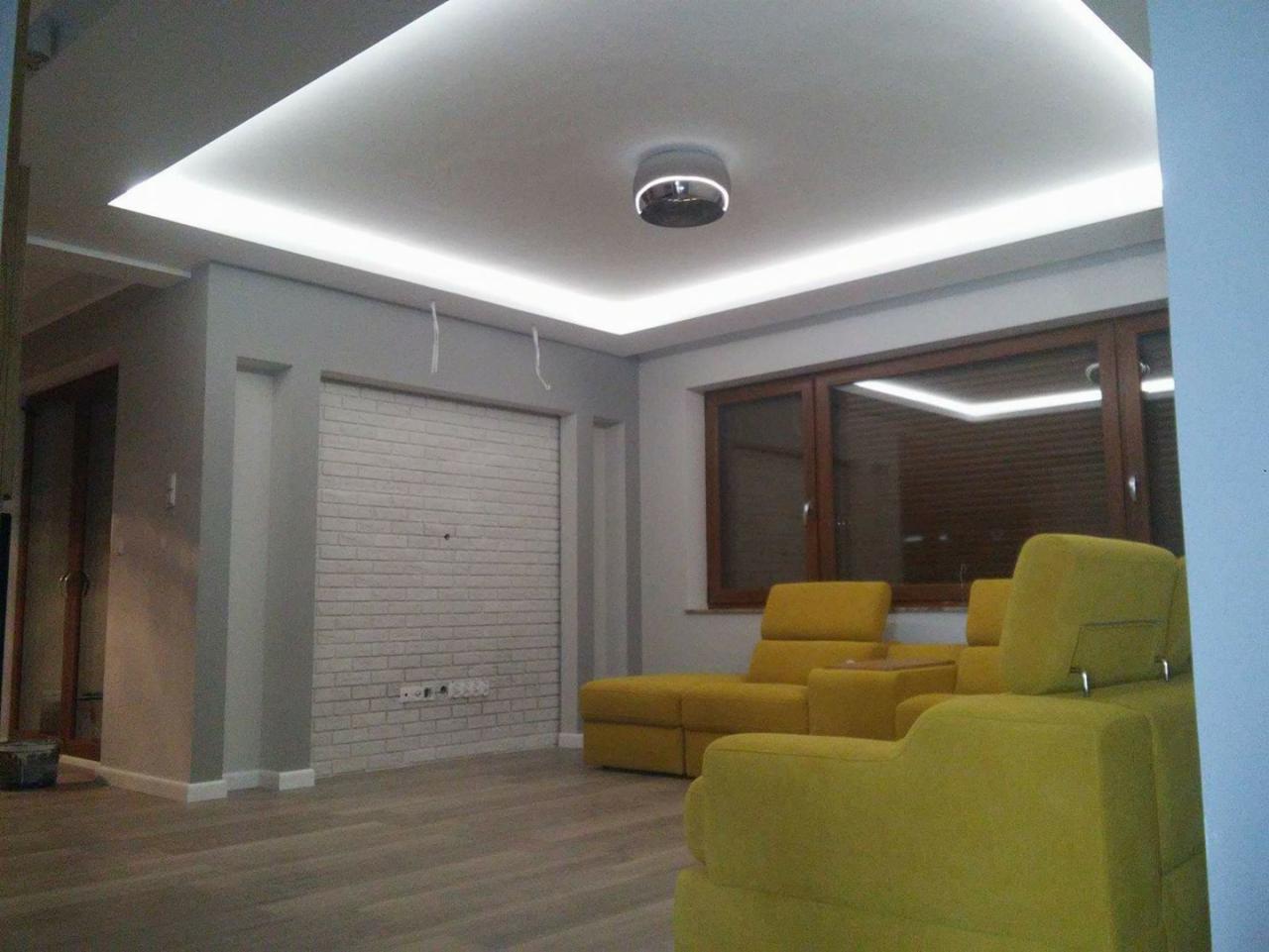 Jasne wnętrze salonu z podświetlanym sufitem kasetonowym LED, szarymi ścianami, imitacją białej cegły na ścianie, drewnianą podłogą i żółtymi meblami wypoczynkowymi.