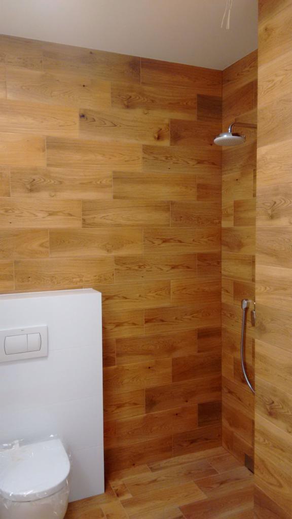Kabina prysznicowa wyłożona płytkami imitującymi drewno, z widoczną deszczownicą i toaletą ze spłuczką podtynkową.