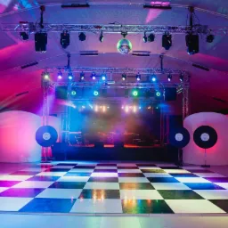 Scena z oświetleniem eventowym, parkietem w szachownicę, kulą dyskotekową, dekoracjami w kształcie płyt winylowych i białymi, podświetlonymi panelami.