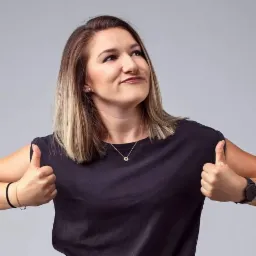 Uśmiechnięta kobieta z blond pasemkami, w czarnej koszulce, pokazuje kciuki w górę na szarym tle. Widoczne kolczyki i zegarek.