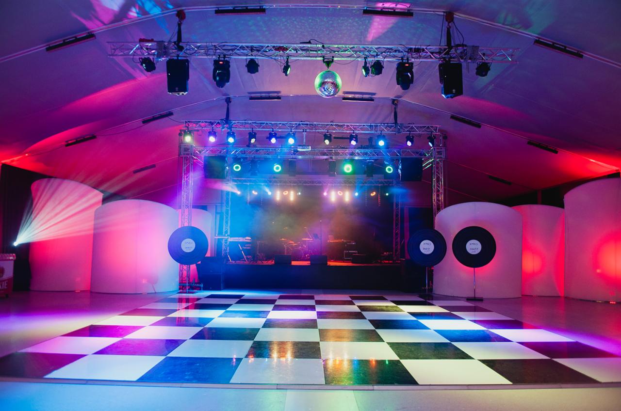 Scena z oświetleniem eventowym, parkietem w szachownicę, kulą dyskotekową, dekoracjami w kształcie płyt winylowych i białymi, podświetlonymi panelami.