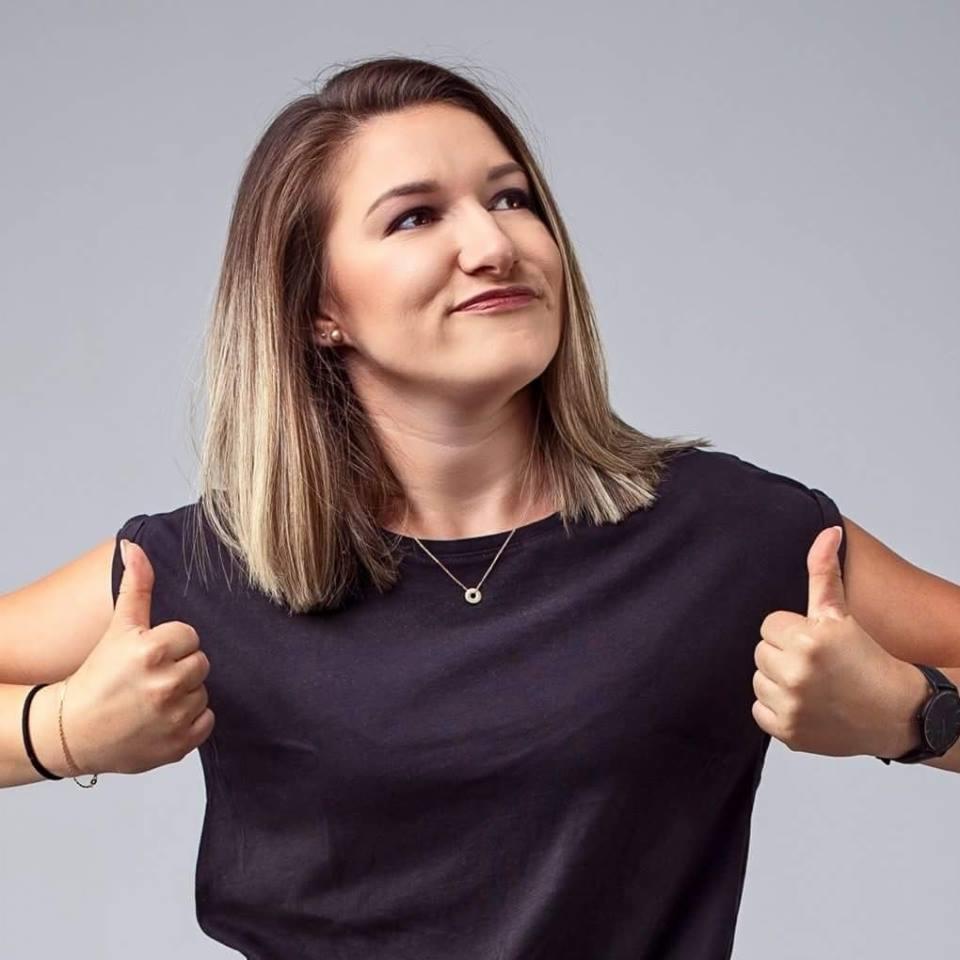 Uśmiechnięta kobieta z blond pasemkami, w czarnej koszulce, pokazuje kciuki w górę na szarym tle. Widoczne kolczyki i zegarek.