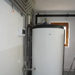 Biały zasobnik ciepłej wody użytkowej marki Buderus z licznymi rurami z izolacją termiczną oraz elementami instalacji, widoczny w pomieszczeniu z białymi kafelkami i drabiną opartą o ścianę.