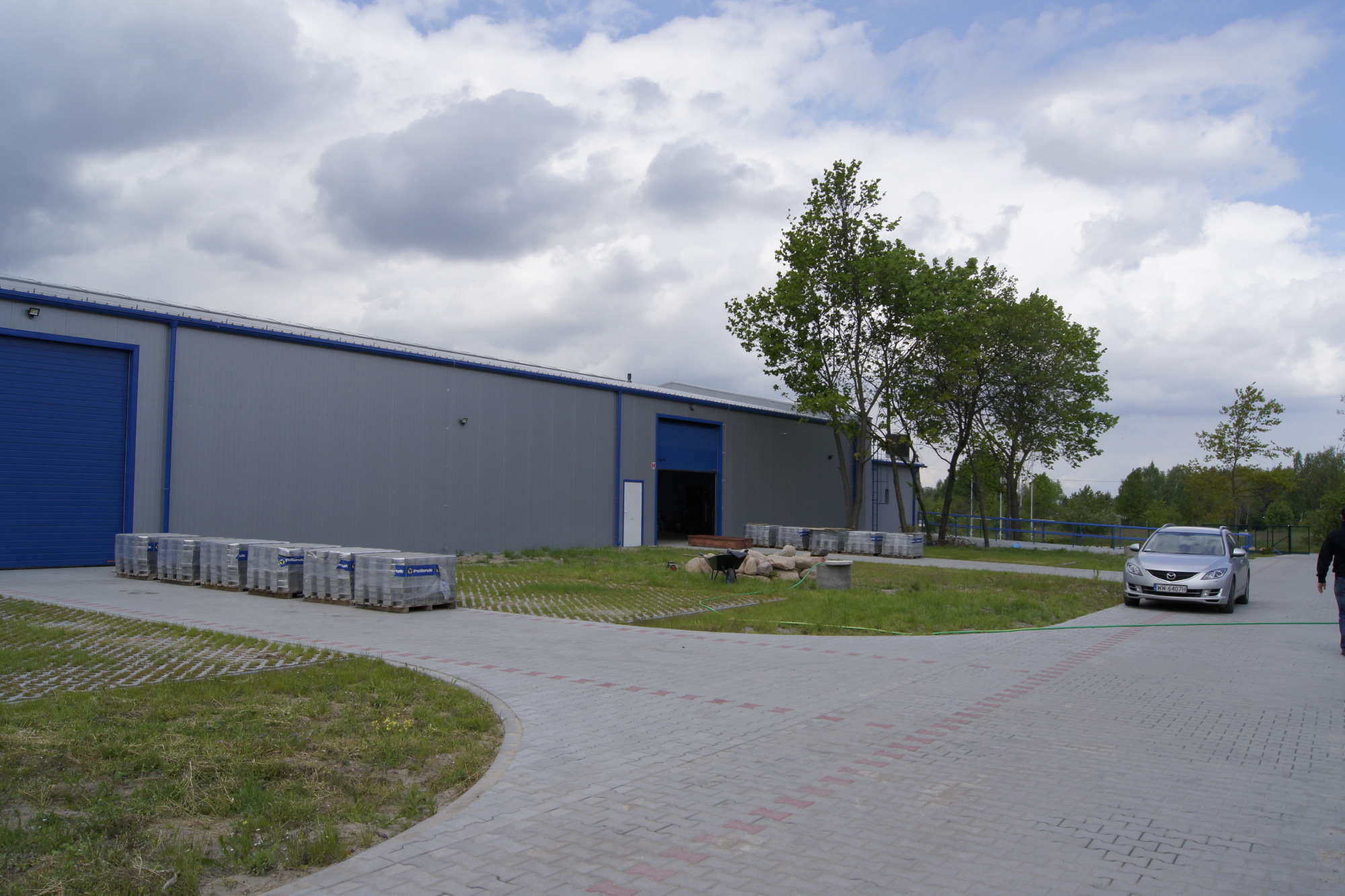 Szary budynek magazynowy z niebieskimi bramami i ułożonymi kostkami brukowymi na paletach przed budynkiem, z widocznym samochodem osobowym na podjeździe.