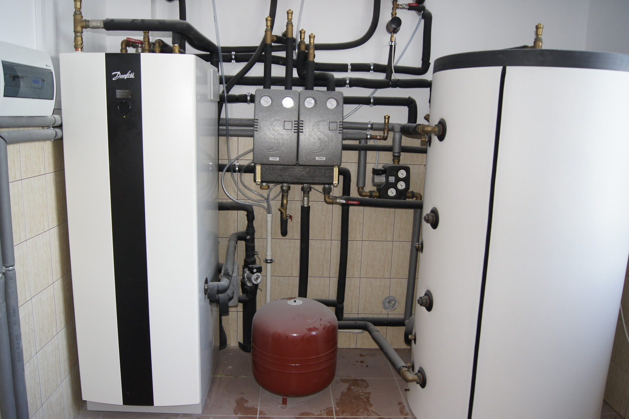 Instalacja grzewcza w pomieszczeniu technicznym z widocznym zbiornikiem wyrównawczym, licznymi rurami i elementami sterującymi, na tle wykafelkowanej ściany.