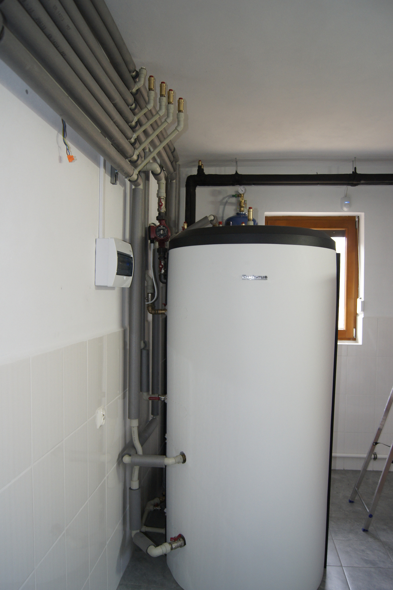 Biały zasobnik ciepłej wody użytkowej marki Buderus z licznymi rurami z izolacją termiczną oraz elementami instalacji, widoczny w pomieszczeniu z białymi kafelkami i drabiną opartą o ścianę.