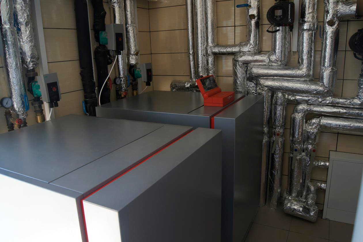 Dwie szare jednostki kotłowe z czerwonymi akcentami, połączone rurami w izolacji aluminiowej, z czerwonym urządzeniem diagnostycznym na górze jednej z jednostek, w kotłowni wyłożonej płytkami.