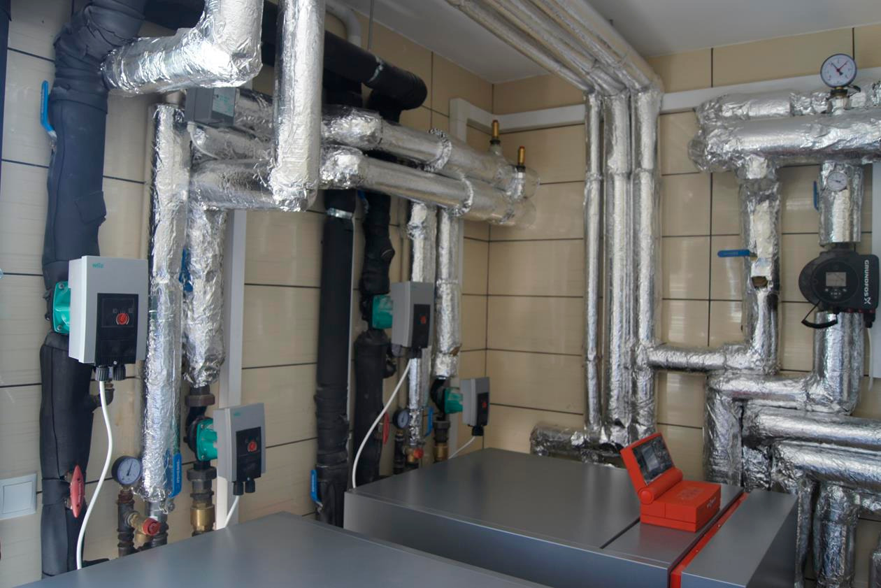 Instalacja z rurami w izolacji aluminiowej, licznymi zaworami i manometrami, widoczne pompy obiegowe z elektronicznymi sterownikami oraz czerwony tester instalacji na metalowej obudowie urządzenia.