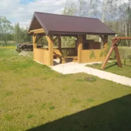 Drewniana altana z brązowym dachem na trawniku, z drewnianą huśtawką w tle, ścieżka z kamienia prowadzi do altany.