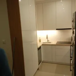 Nowoczesna, jasna kuchnia z białymi, lakierowanymi frontami szafek, drewnianym akcentem na boku wysokiej zabudowy, oświetleniem podszafkowym i błyszczącą, kremową podłogą.