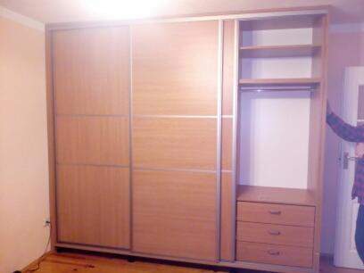 Jasna szafa przesuwna z półkami i szufladami, widoczna w części pokoju z drewnianą podłogą i fragmentem osoby w koszuli w kratę przy drzwiach po prawej stronie.