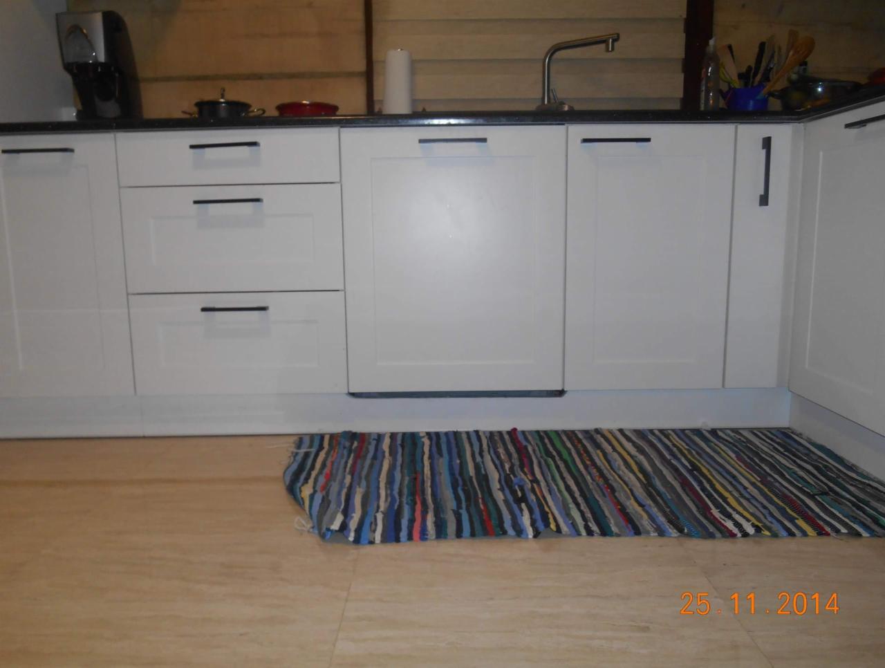 Biała zabudowa kuchenna z czarnym blatem i prostymi, podłużnymi uchwytami, uzupełniona o kolorowy, pleciony chodnik na podłodze.