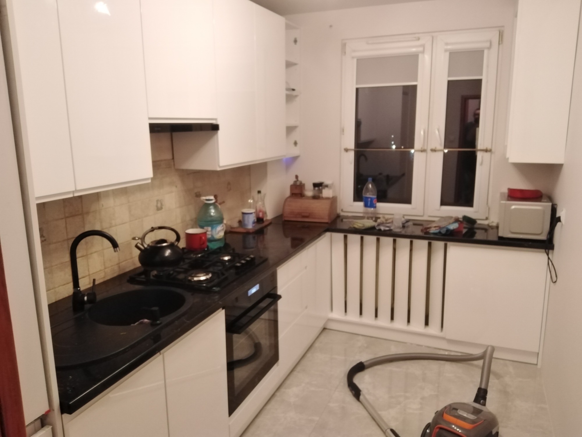 Biała kuchnia z czarnym blatem, widoczny zlewozmywak, płyta gazowa, piekarnik oraz odkurzacz na podłodze.