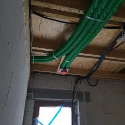 Zielone, karbowane rury wentylacyjne przymocowane metalowymi obejmami do drewnianego stropu w surowym stanie, widoczne także czarne kable elektryczne, ściany z betonu komórkowego i okno...