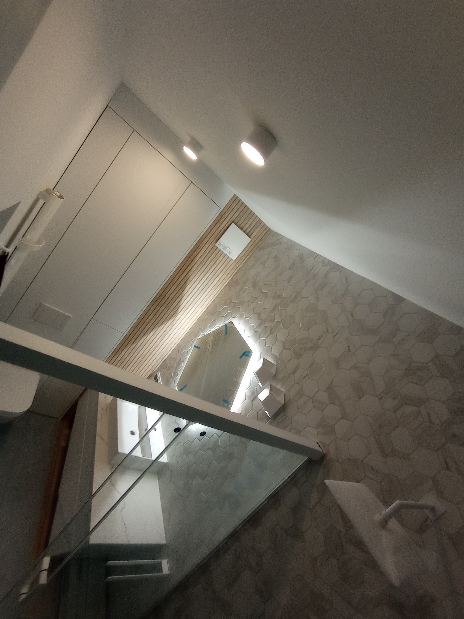 Nowoczesna łazienka z heksagonalnymi płytkami na ścianie, kabiną prysznicową ze szklanymi drzwiami i drewnianymi panelami dekoracyjnymi pod sufitem, oświetlona punktowymi lampami.