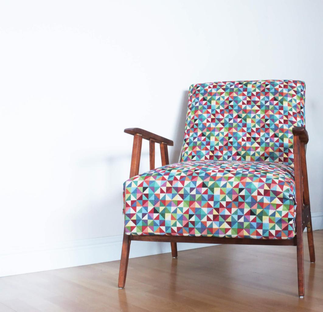 Retro fotel z drewnianymi podłokietnikami i tapicerką w kolorowe trójkąty na tle białej ściany.