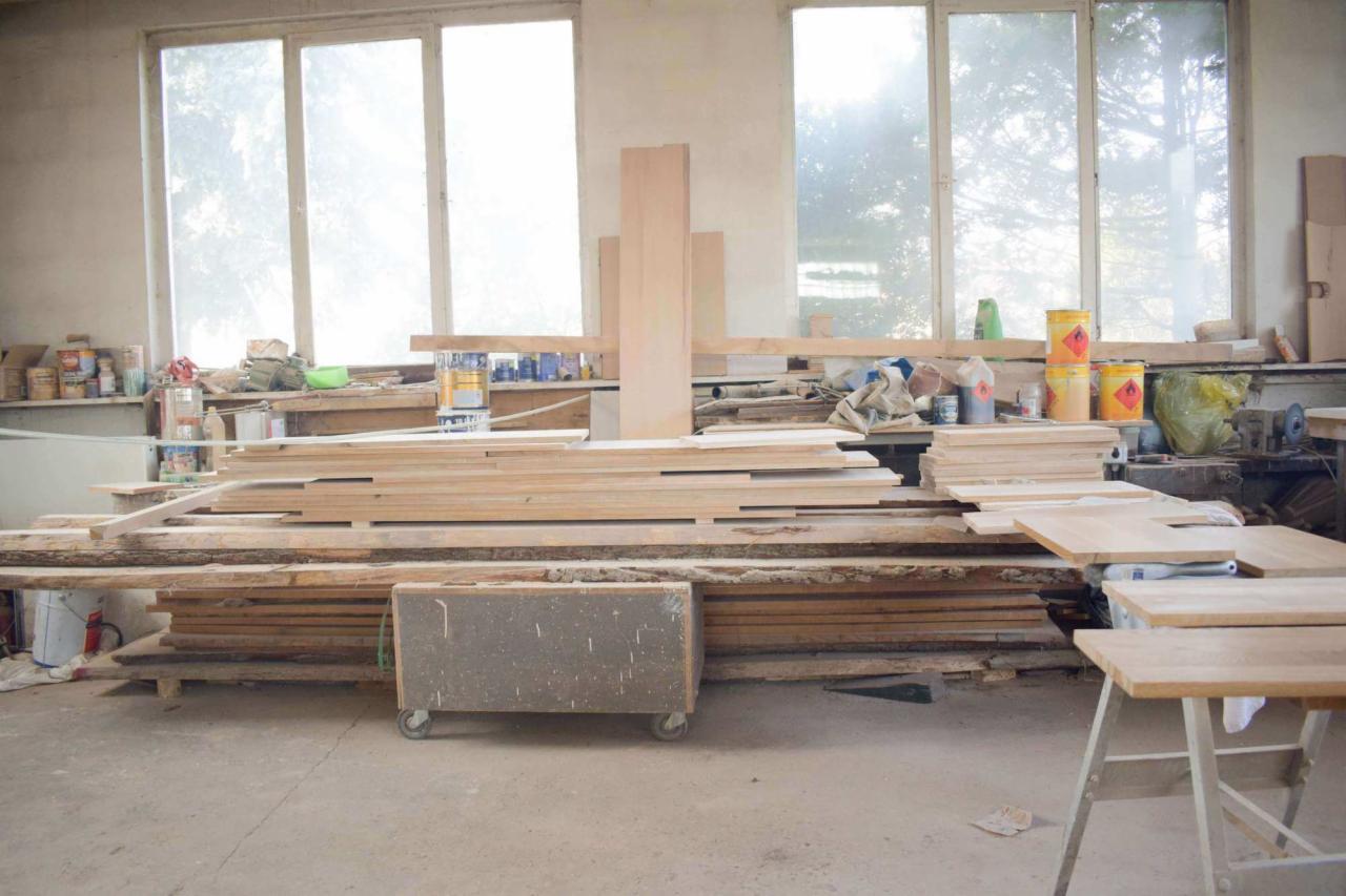 Warsztat stolarski: Stosy surowych desek i blatów drewnianych pod oknem, widoczne farby i narzędzia na półkach, w tle maszyna stolarska.