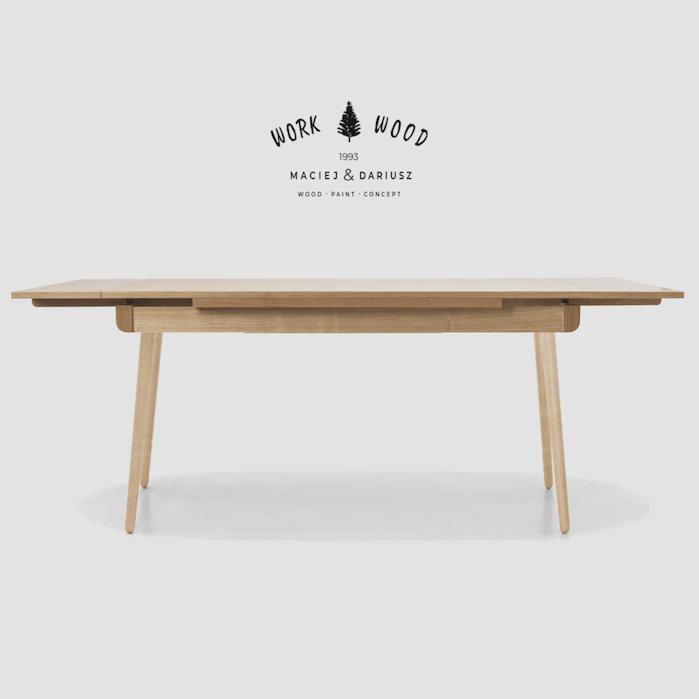 Rozkładany stół z jasnego drewna na tle szarego studia, z widocznym logo producenta 'Work Wood' w górnej części zdjęcia.