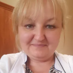 Portret uśmiechniętej kobiety w białym fartuchu lekarskim, z blond włosami i zielonymi oczami, na rozmytym tle ściany i drewnianej szafki.